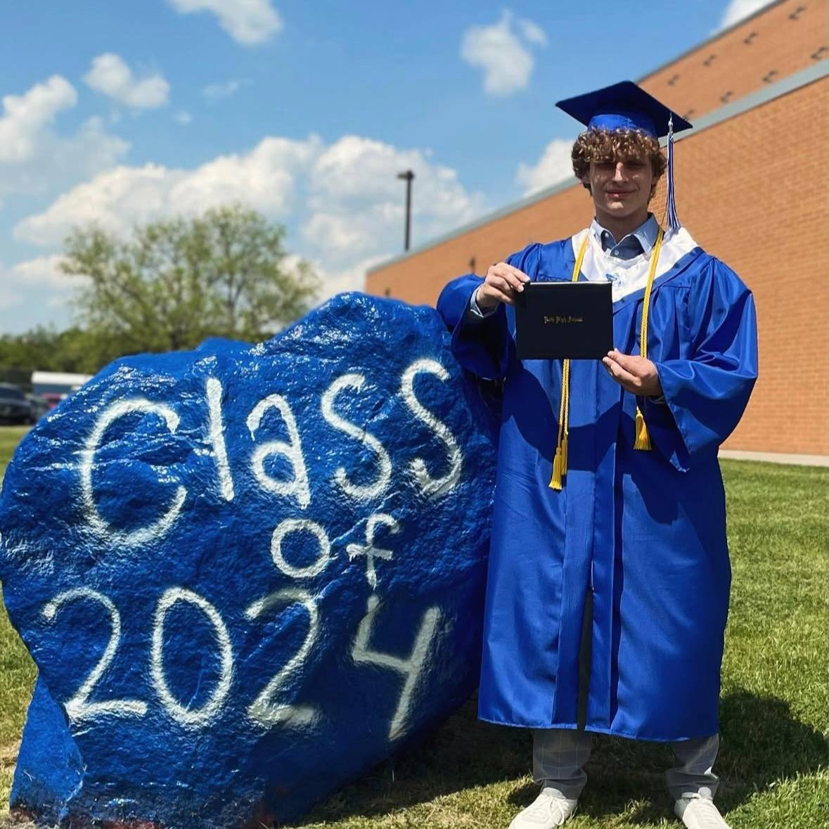 student wearing graduation robes holding a diploma next to a rock with 2024 written on it